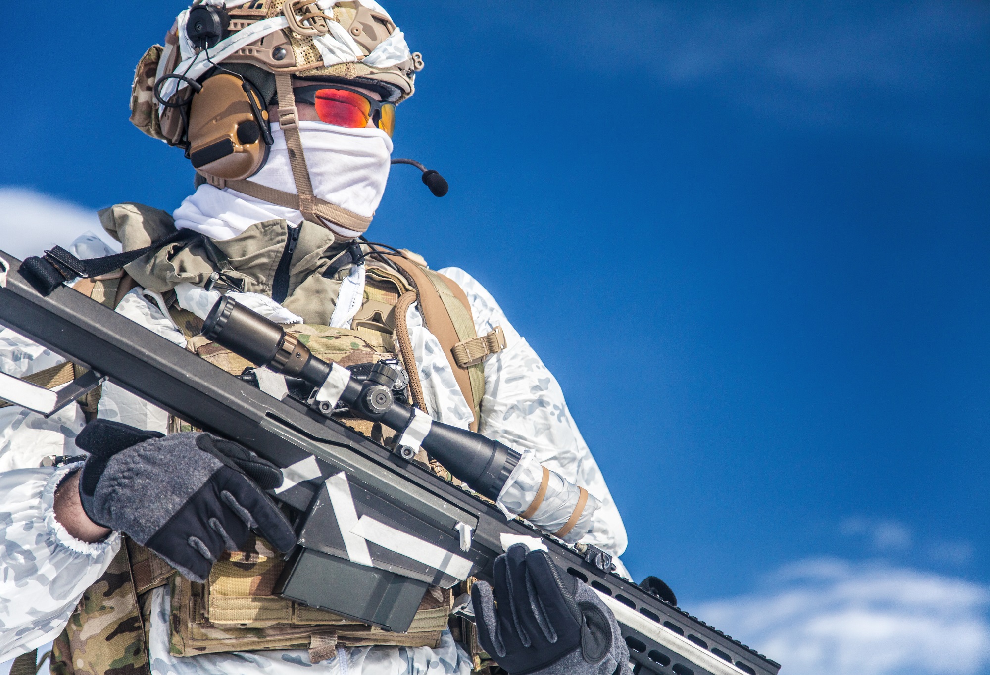 Army serviceman in winter camo somewhere in the Arctic. He wears chest rig and huge backpack, suffers from the extreme cold and strong wind, but endures while mission continues. Blue sky background